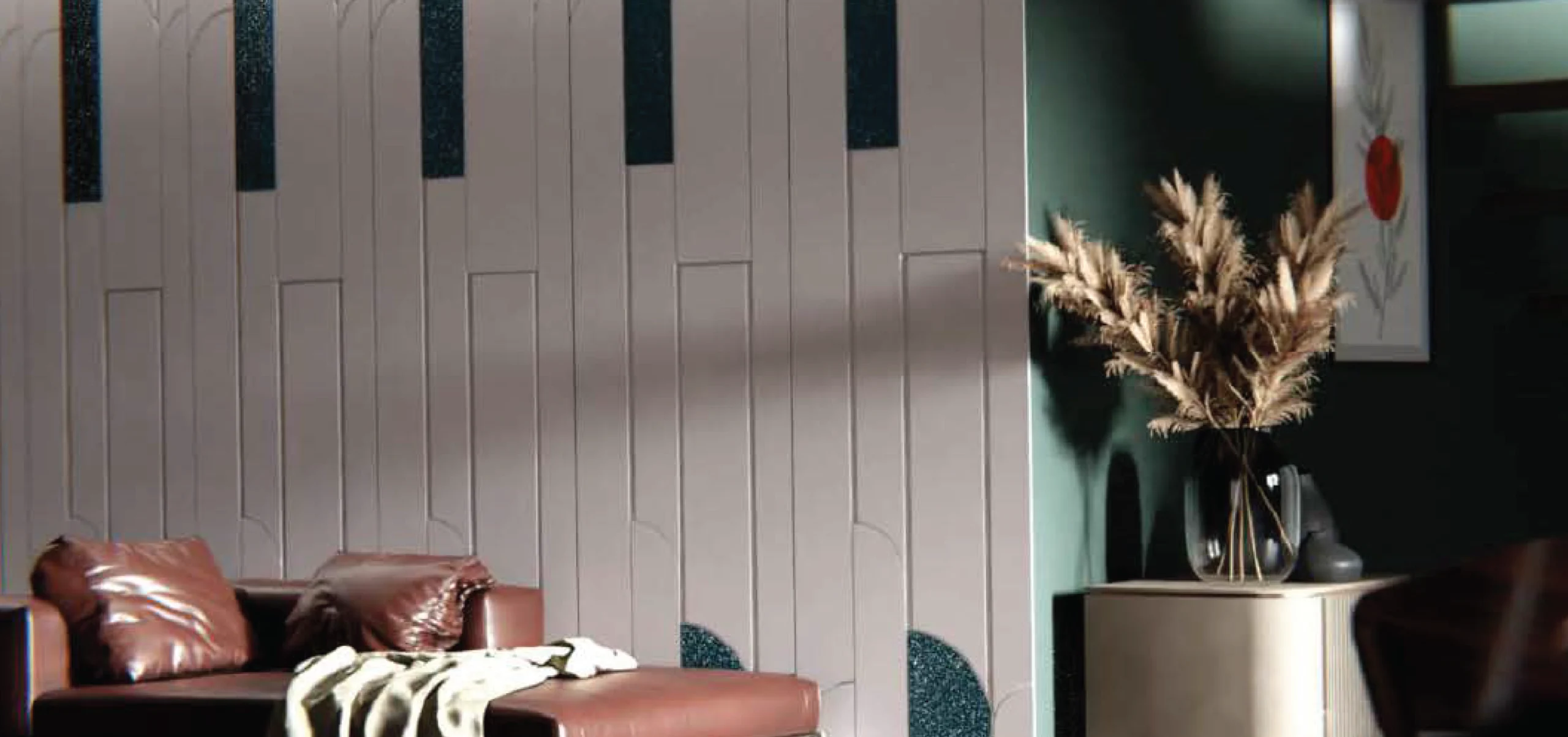 A cozy modern living room corner featuring a brown leather sofa with pillows, a textured beige wall with vertical panel details and dark blue accents, a green wall with a framed flower artwork, and a decorative vase with dried pampas grass on a cream console table.