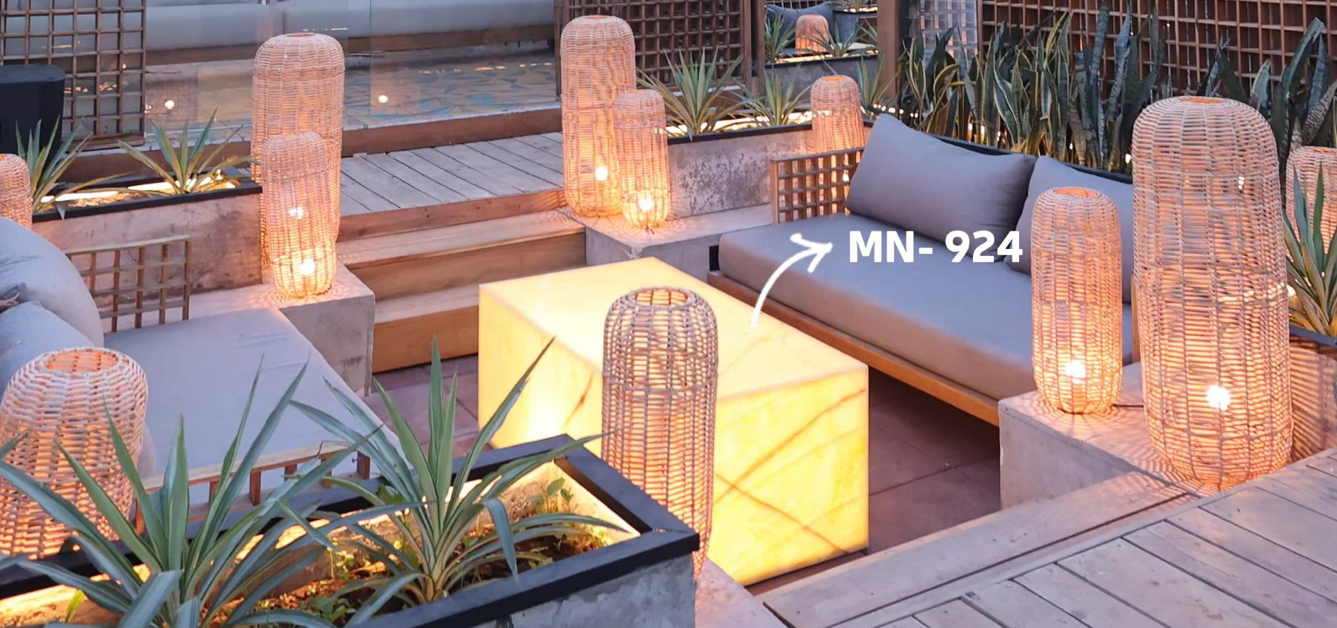 A cozy outdoor seating area featuring a glowing alabaster table (model MN-924) surrounded by woven rattan lamps, concrete planters with green plants, and modern gray seating.